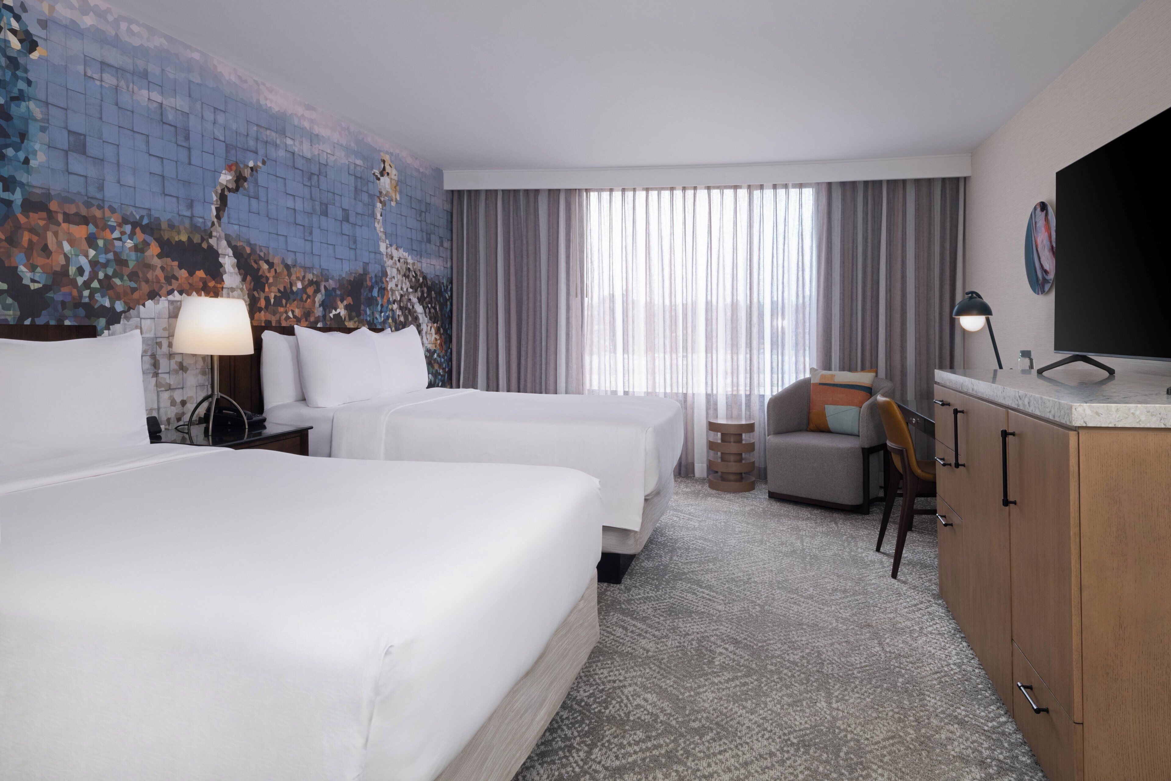A hotel room with two beds, a desk, a chair, a TV, and a large window with curtains.