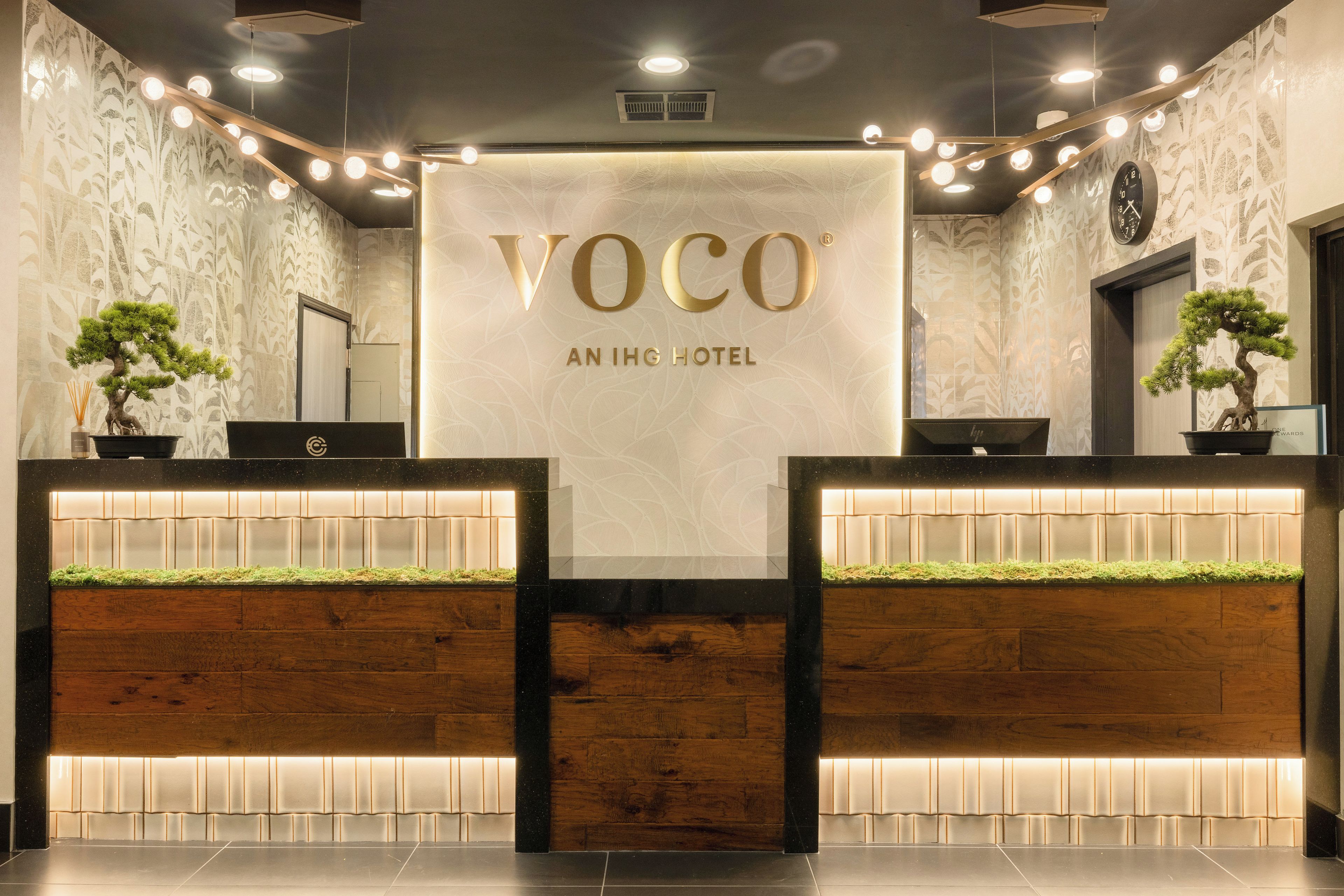 A reception desk with a sign reading 'VOCO AN IHG HOTEL' and two potted plants.