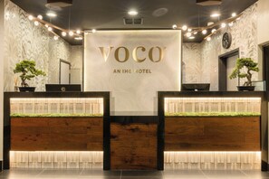 Un comptoir d’accueil avec une enseigne indiquant « VOCO, UN HÔTEL IHG » et deux plantes en pot.