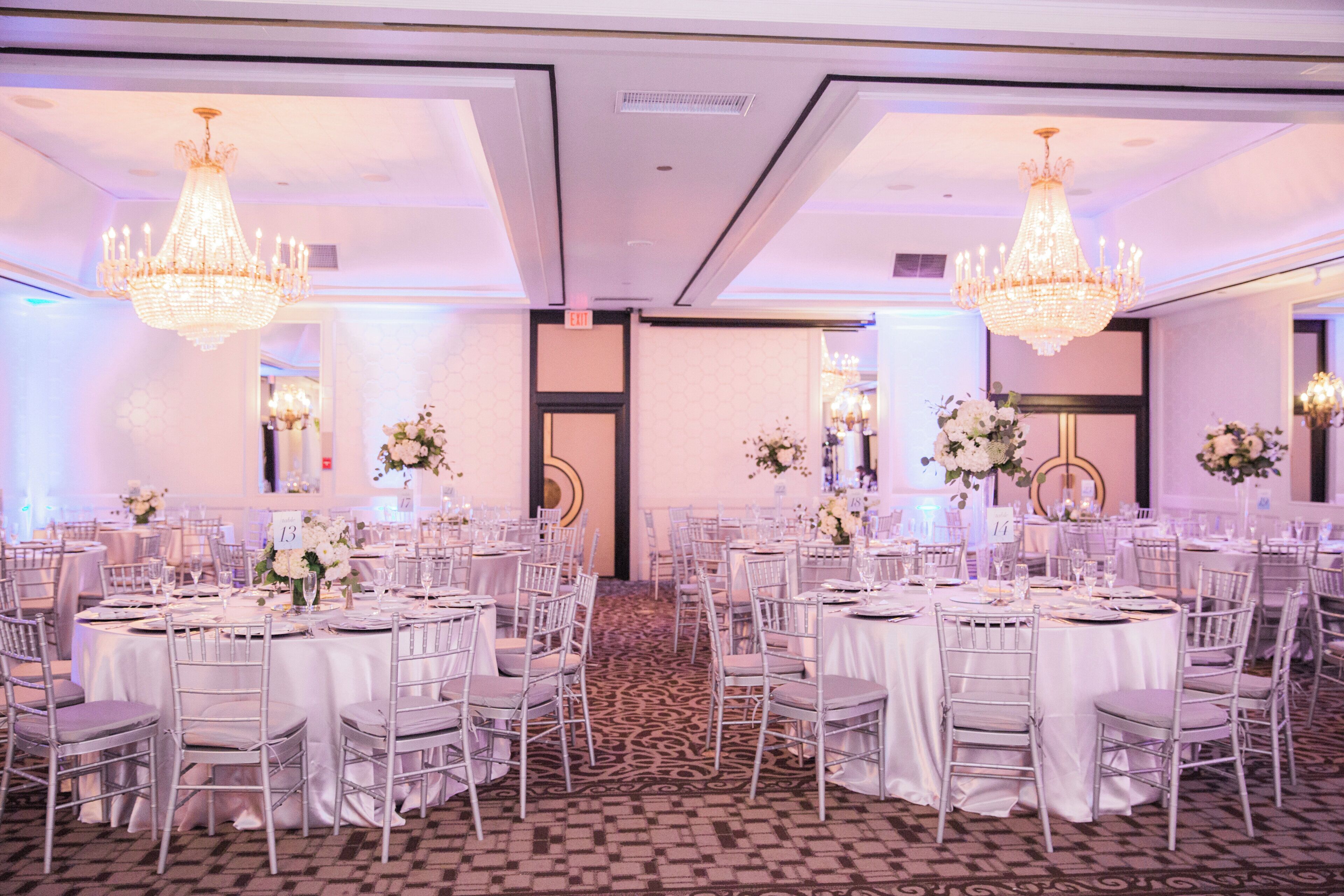 Un salón de banquetes con mesas redondas, manteles blancos y sillas plateadas.
