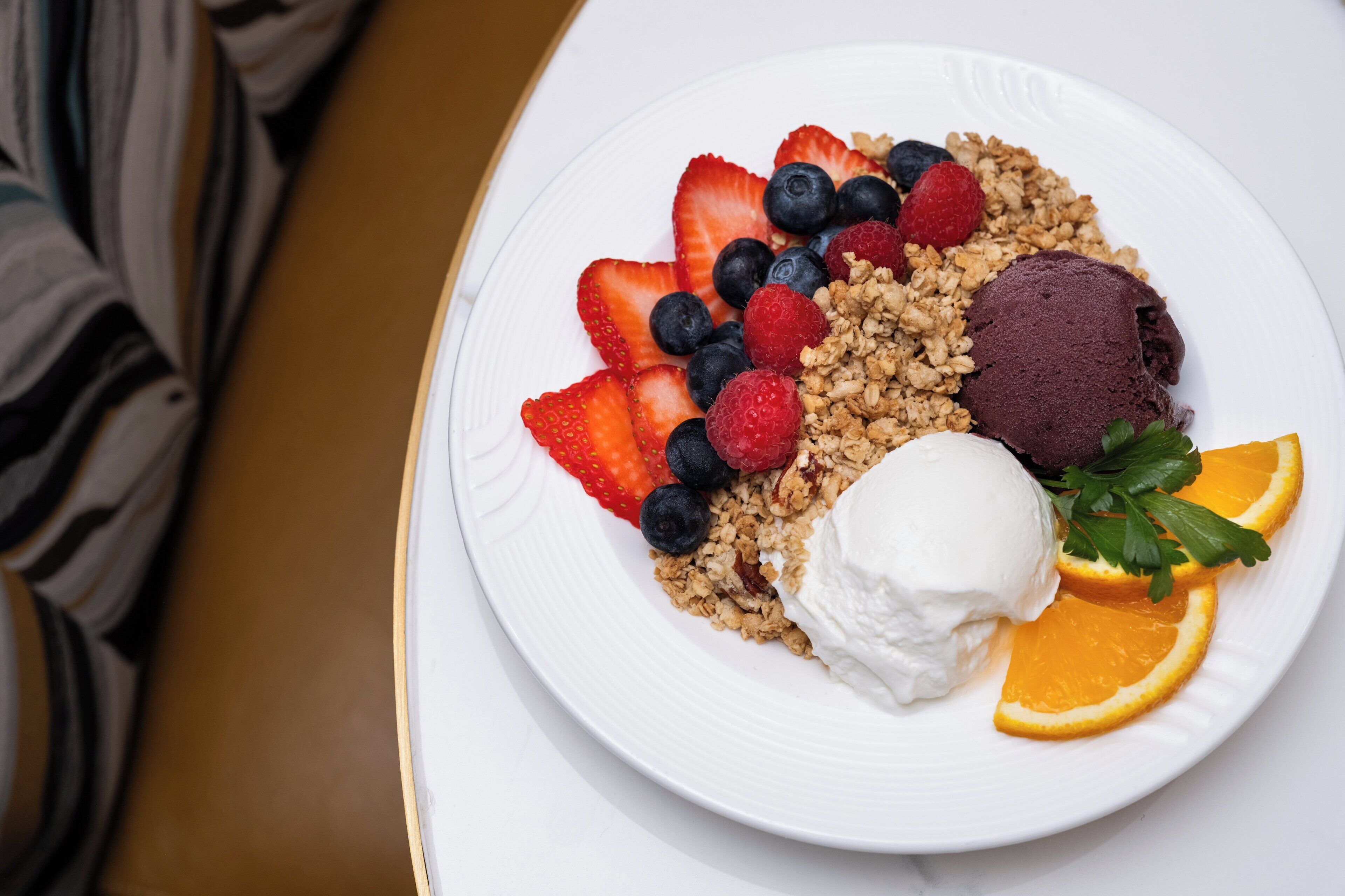 Un plato con un postre de granola, bayas, una porción de helado y una rodaja de naranja.