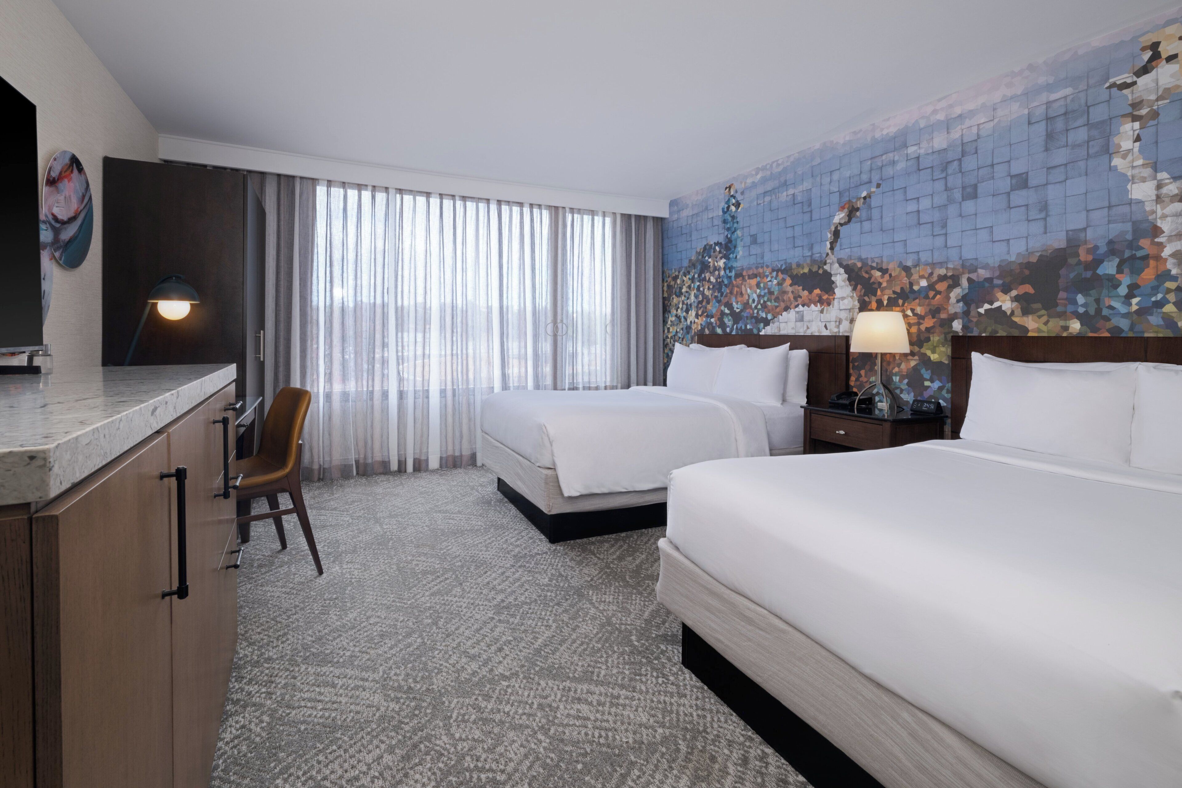 Una habitación de hotel con cama, escritorio, silla, lámpara y un ventanal con cortinas.