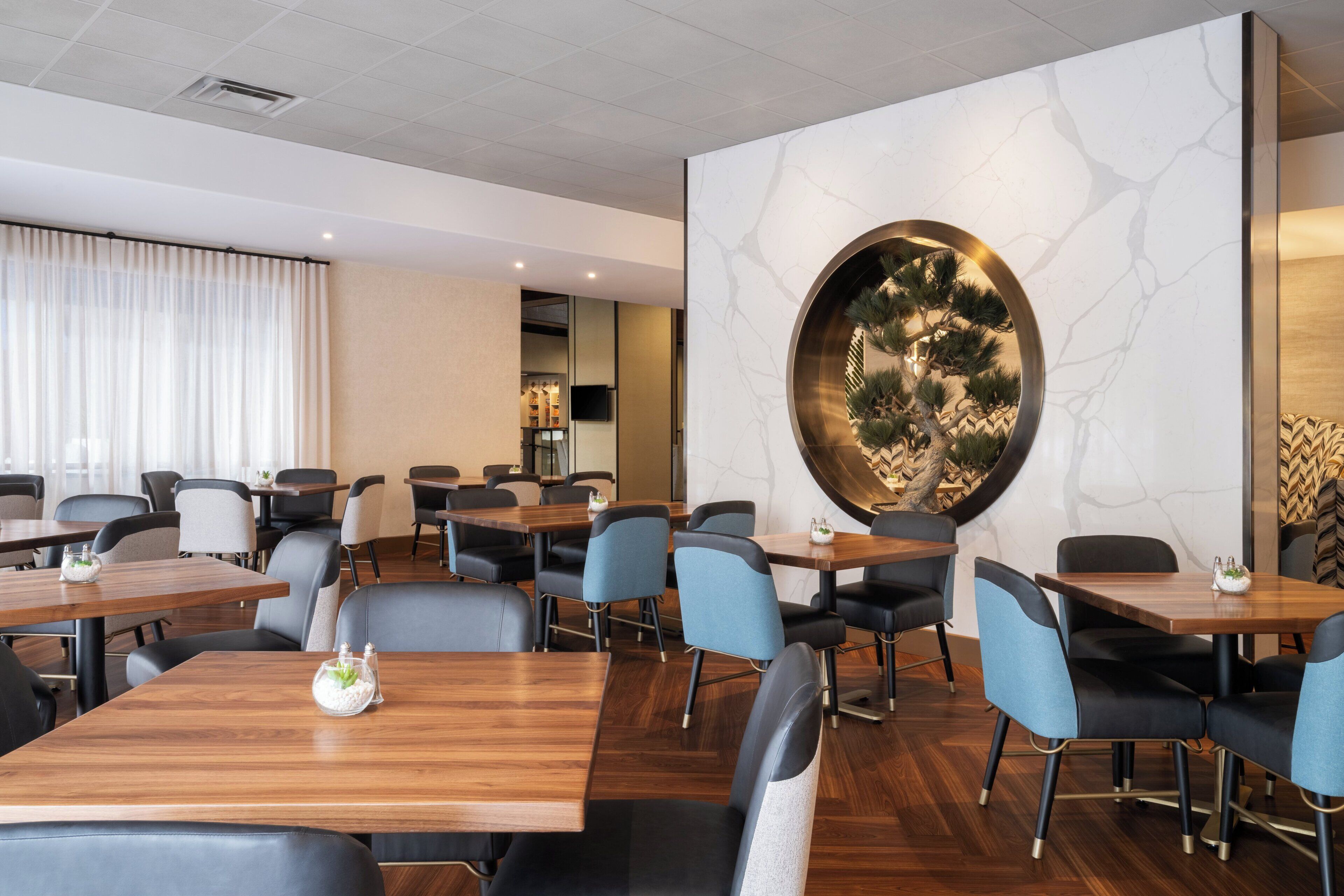 Un comedor moderno con mesas y sillas de madera, un gran elemento decorativo en la pared y vistas a la cocina al fondo.