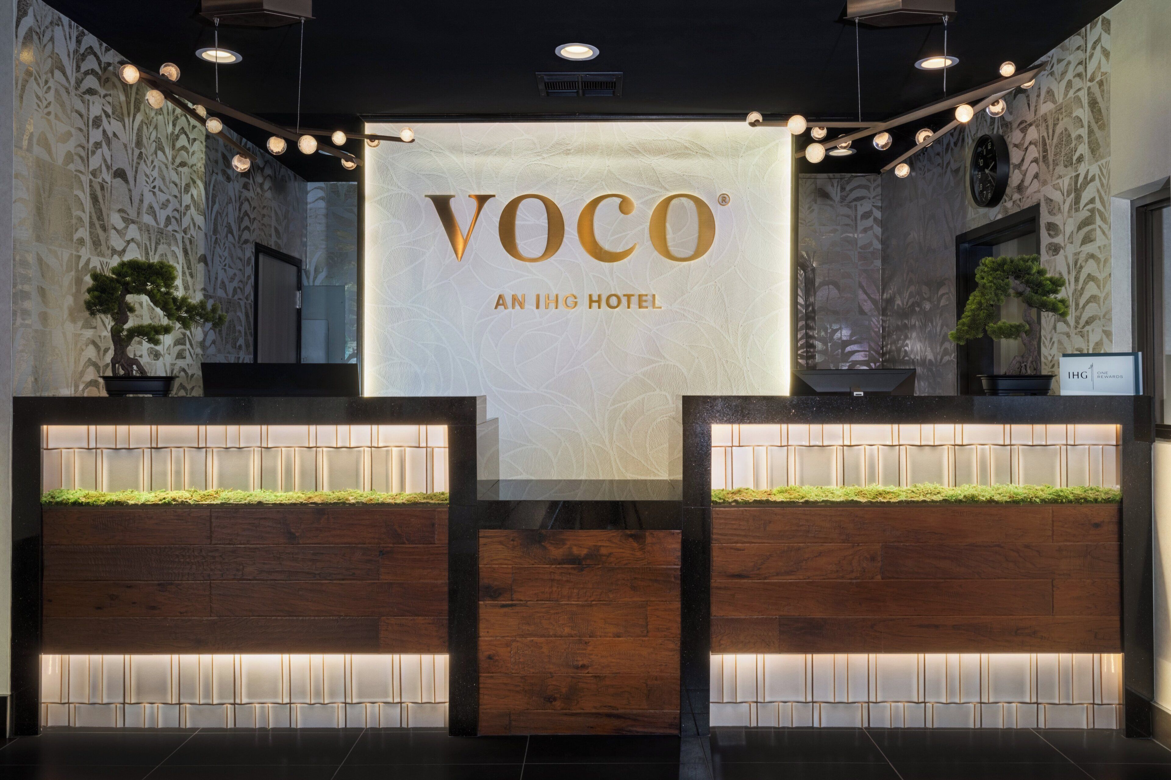 A reception desk with a sign reading 'VOCO AN IHG HOTEL' and two potted plants.