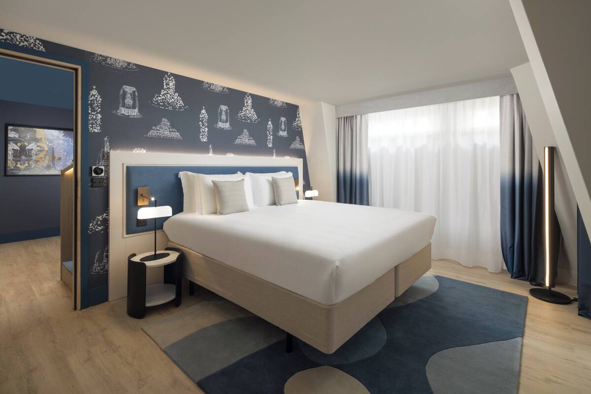Een moderne hotelkamer met een groot bed, een nachtkastje met lamp, een wandklok en een groot raam met gordijnen.