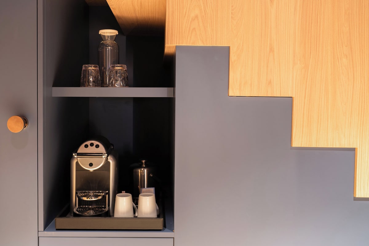A kitchenette with a coffee machine, two white mugs, and a glass container on a shelf.