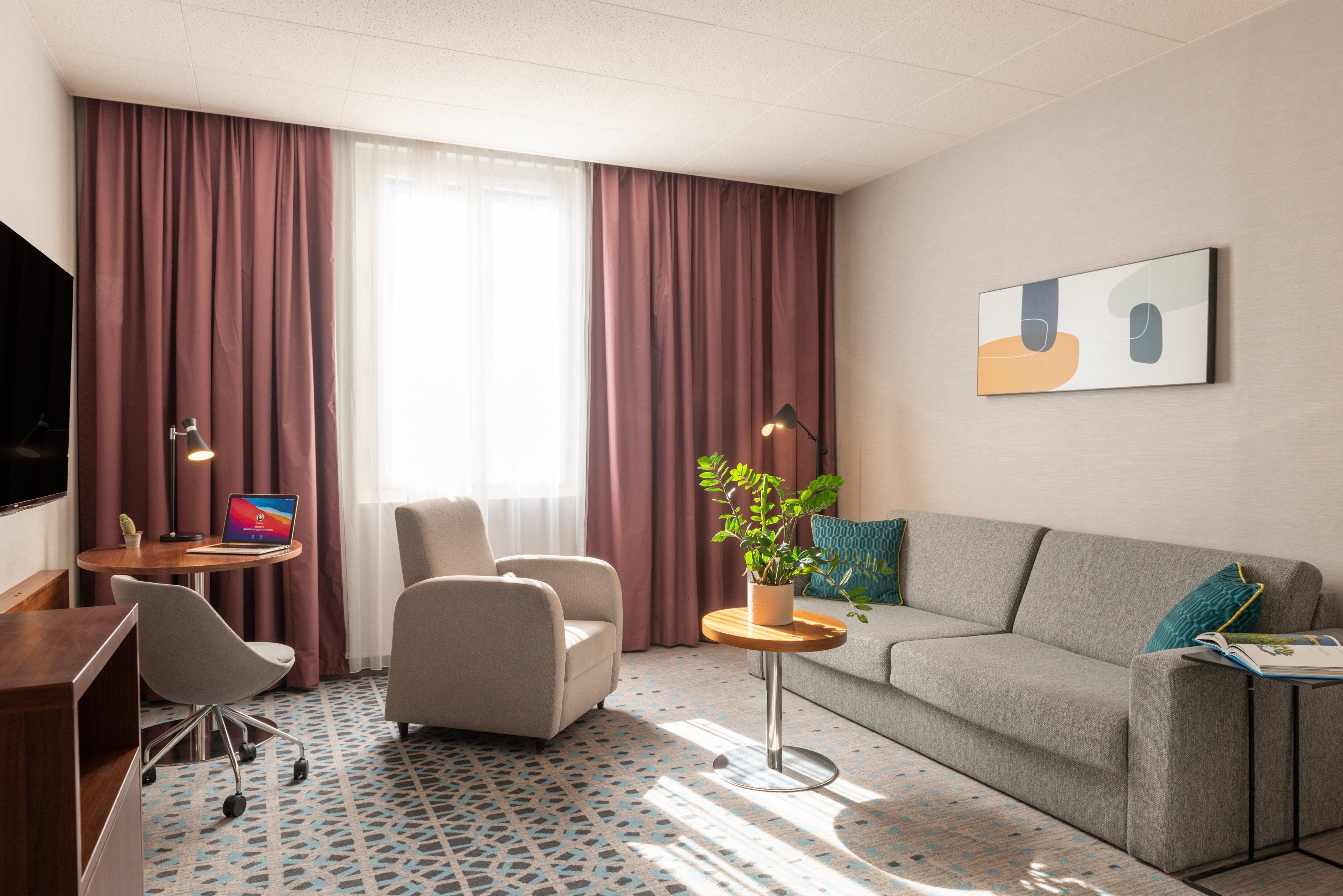 A hotel room with a sofa, armchair, desk, and a potted plant.