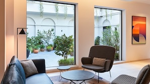 A modern interior with a sofa, armchair, and small table, large windows with plants, and a geometric painting. - Crowne Plaza Paris Republique by IHG (Paris)