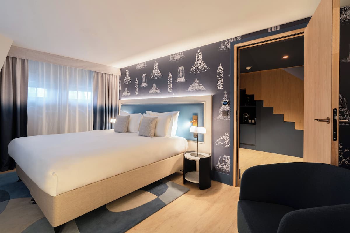 Ett modernt hotellrum med en stor säng, ett litet sidobord, en stol och en trappa som leder upp till våningen ovanför.
