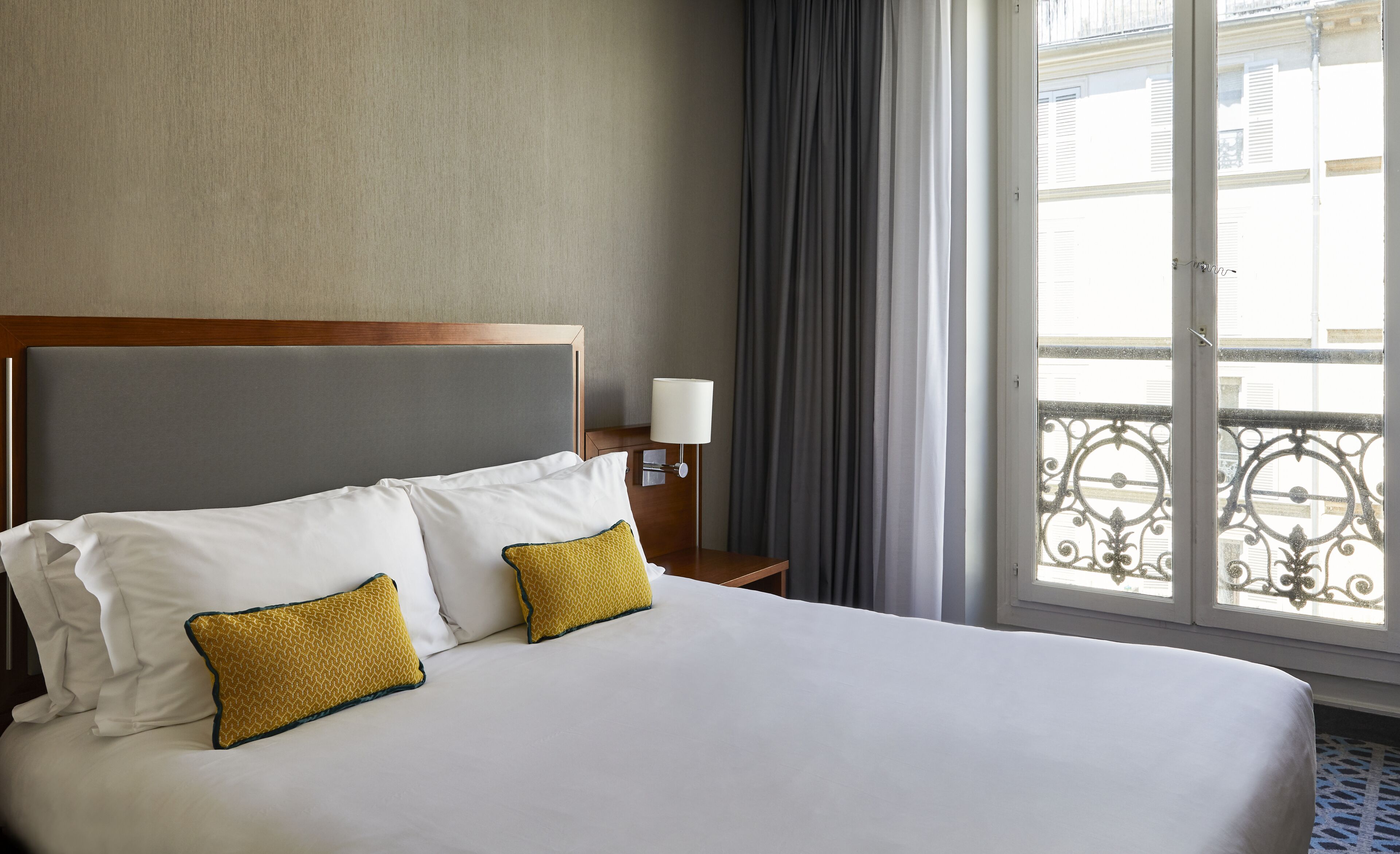 A hotel room with a large bed, two pillows, a nightstand with a lamp, and a view of a balcony with a railing.