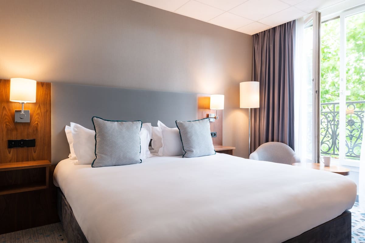 Ett hotellrum med en stor säng, två sängbord och en stol. Det finns två väggmonterade lampor och ett fönster med gardiner.
