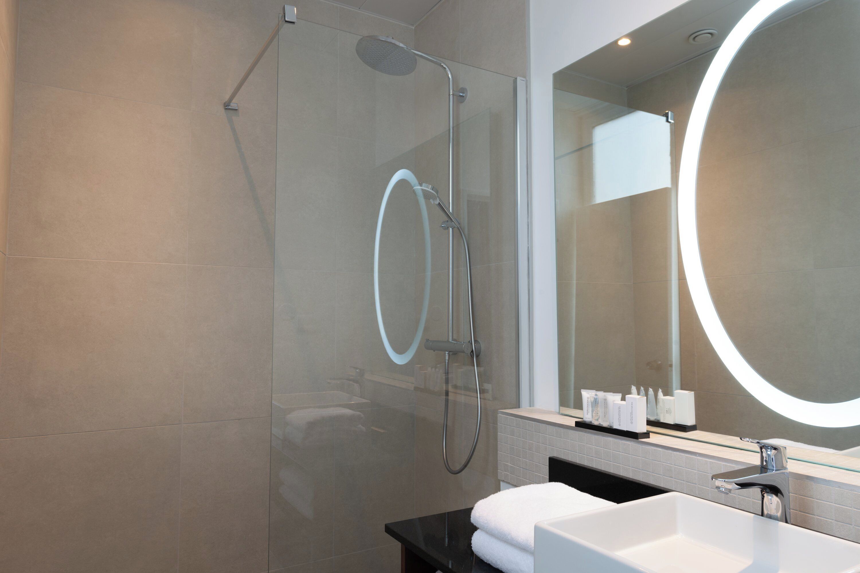 A modern bathroom with a glass-enclosed shower, a large mirror with a circular light, and a black countertop with a white sink.