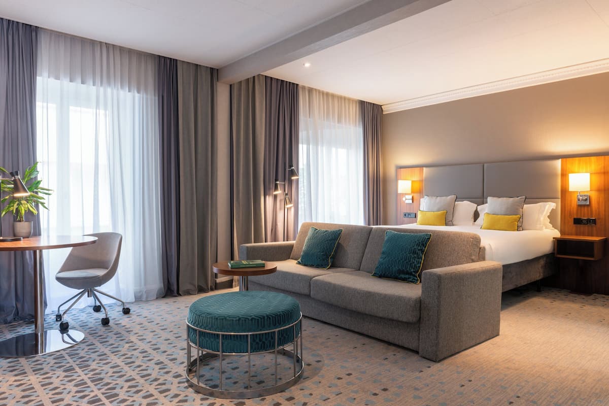 Ett hotellrum med en soffa, en pall och en säng med kuddar. Det finns ett skrivbord med en stol och en lampa.