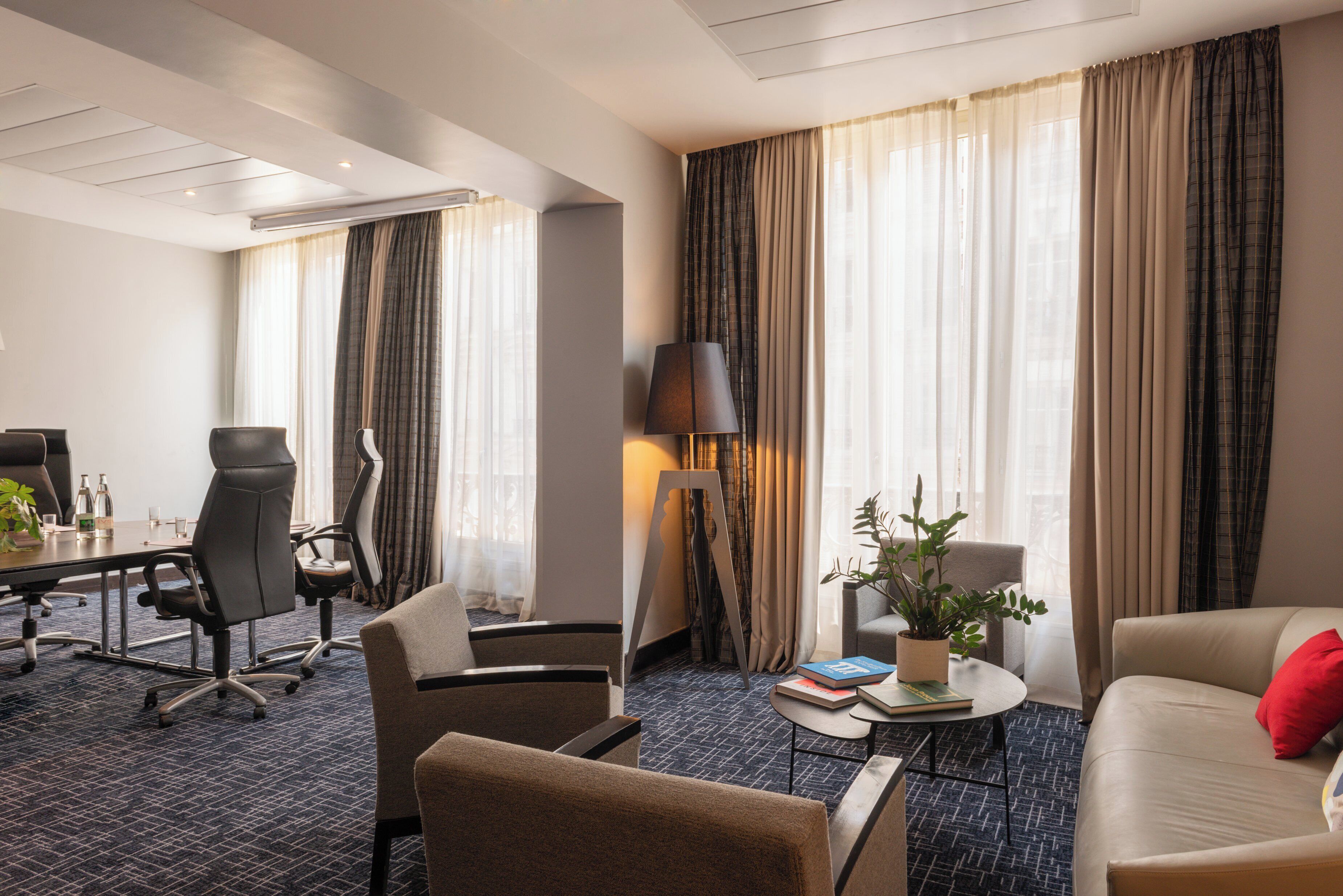 Een hotelkamer met een vergadertafel, bureaustoelen, een bank, een salontafel met boeken en een potplant.