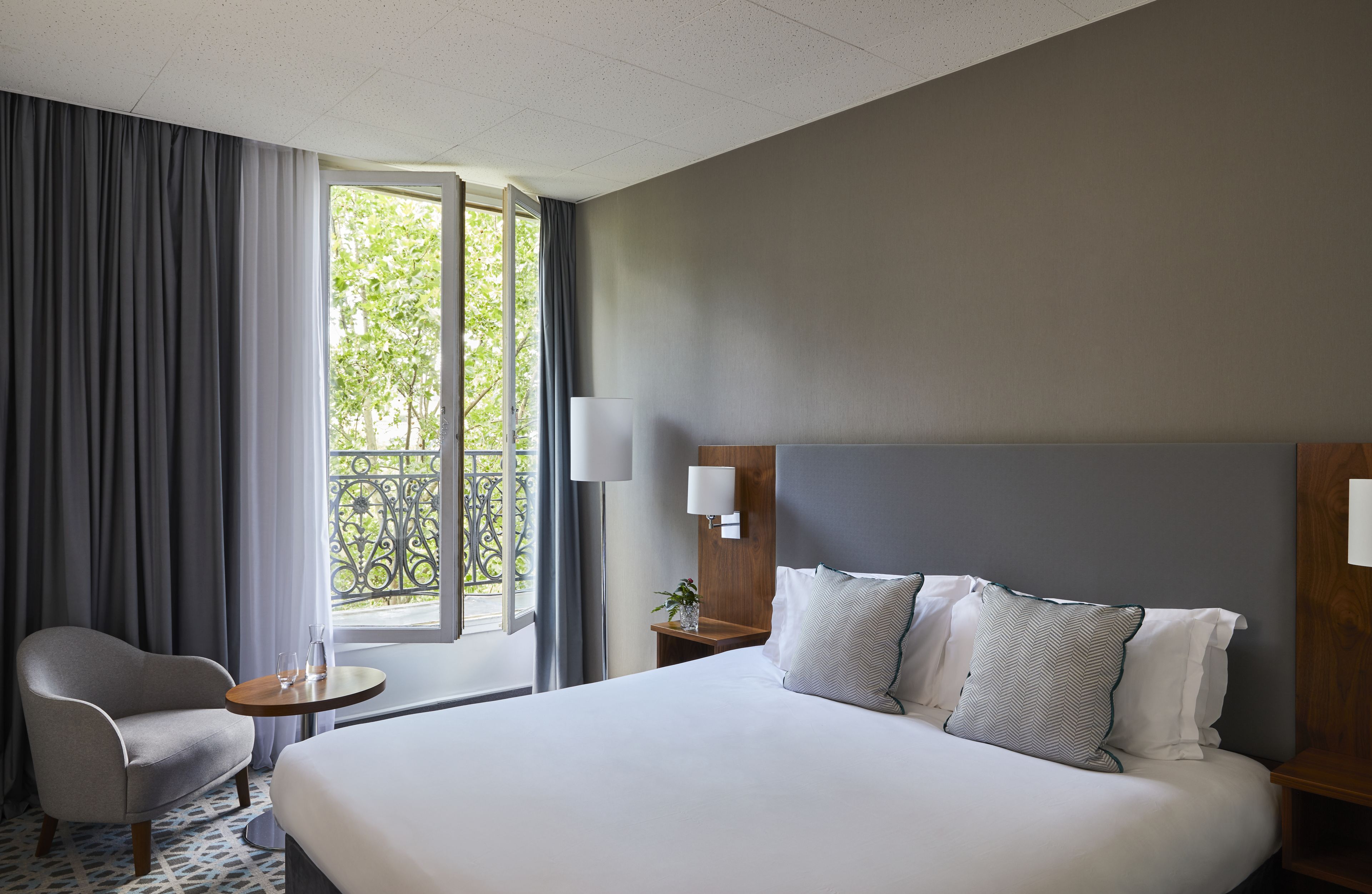 Habitació d'hotel amb un llit gran, una cadira, una taula petita i una finestra amb cortines.
