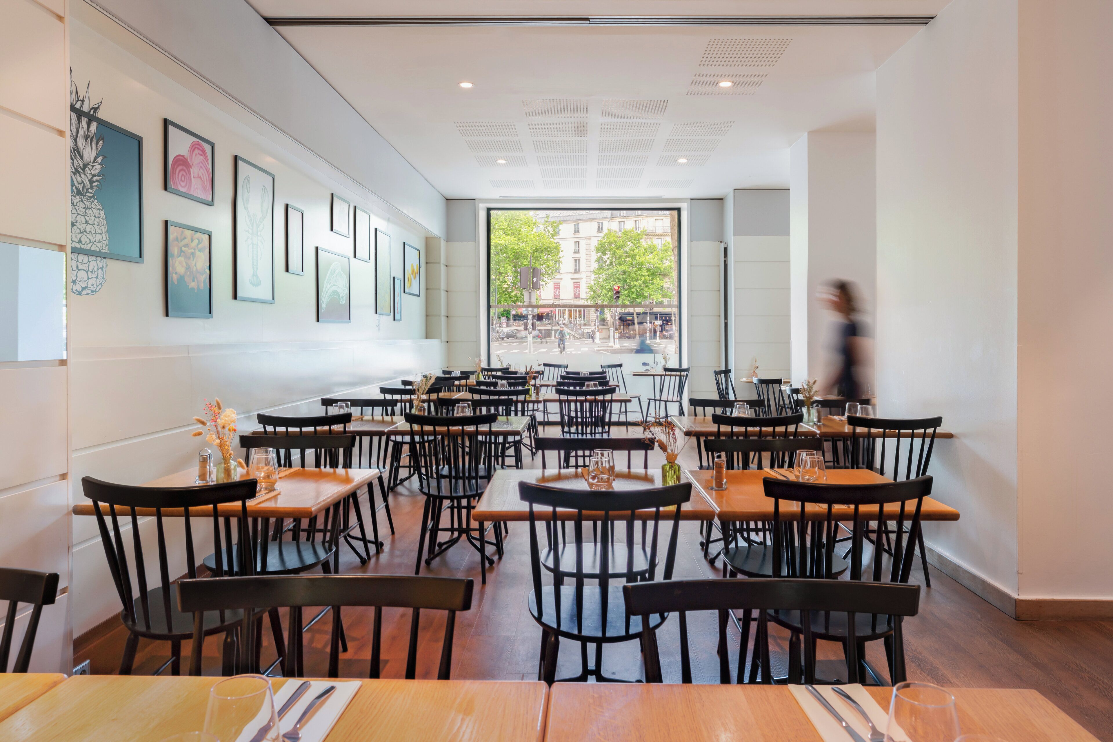 A restaurant interior with multiple tables and chairs, framed artwork on the walls, and a view of the street outside.