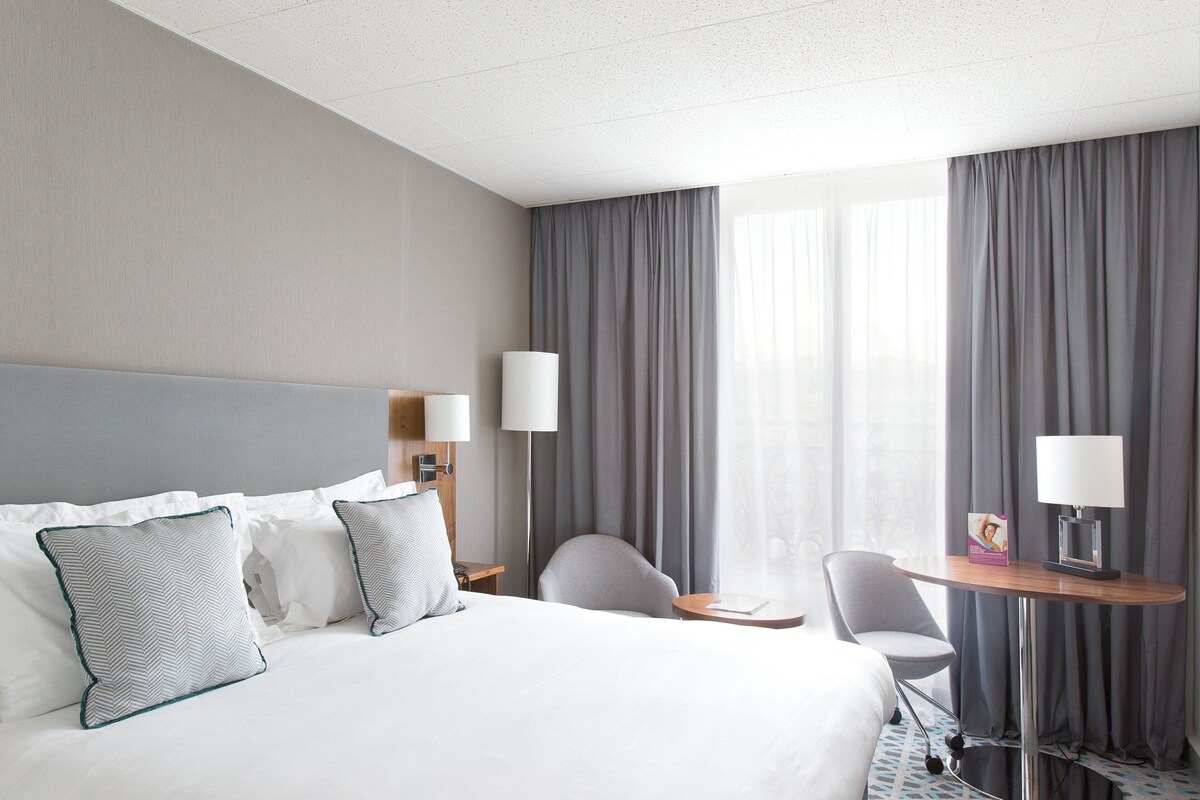 A hotel room with a large bed, two bedside tables, a desk, a chair, and a large window with curtains.