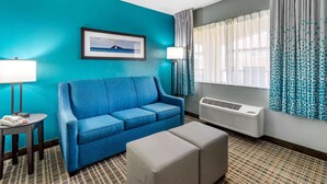 A hotel room with a blue sofa, a small table with a phone, a lamp, and a window with patterned curtains. - Comfort Inn Sun City Center - Ruskin - Tampa South (Sun City Center)