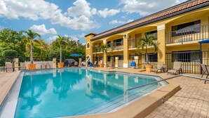 A swimming pool with a tiled deck, surrounded by lounge chairs and umbrellas, and a building with balconies and a tiled roof. - Comfort Inn Sun City Center - Ruskin - Tampa South (Sun City Center)