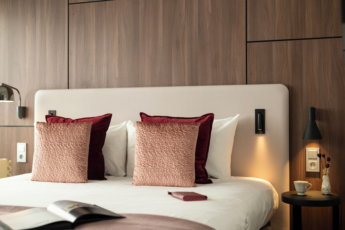 A neatly made bed with two pillows, a bedside table with a cup and a vase, and a wall-mounted light.