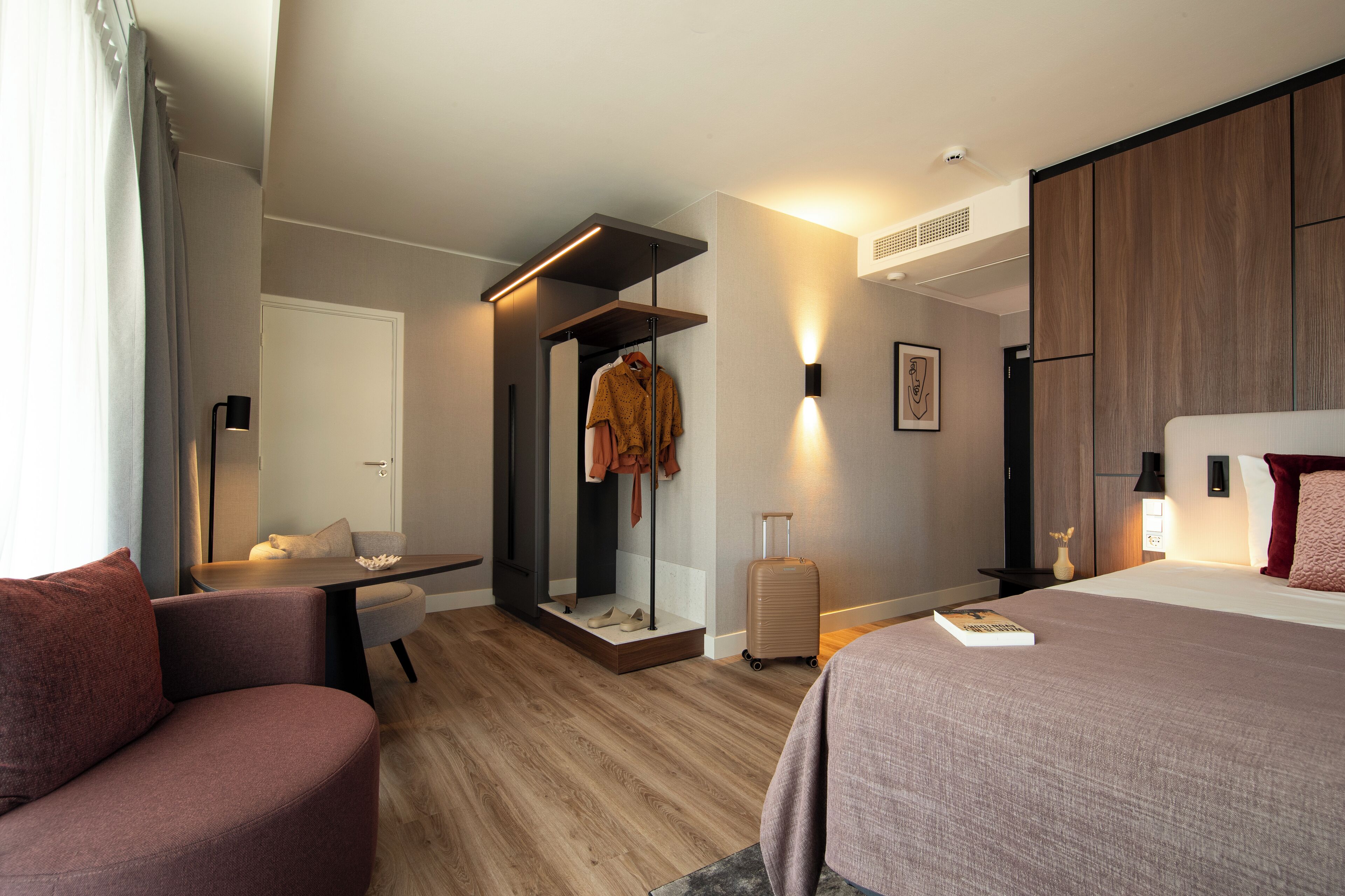 Una habitación de hotel moderna con piso de madera, una cama con almohada roja, un escritorio con silla, un armario y una maleta.