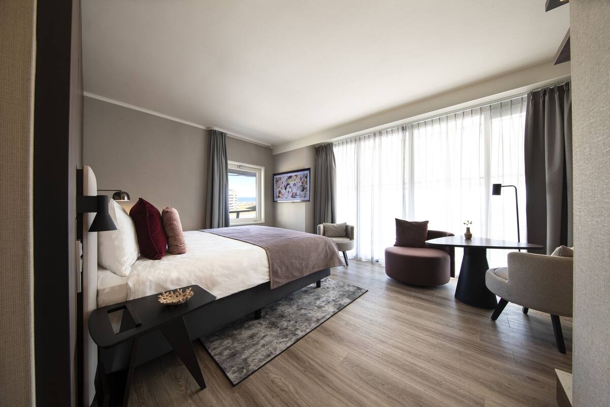 Een moderne hotelkamer met een bed, een bureau, een stoel, een bank en een groot raam met gordijnen.