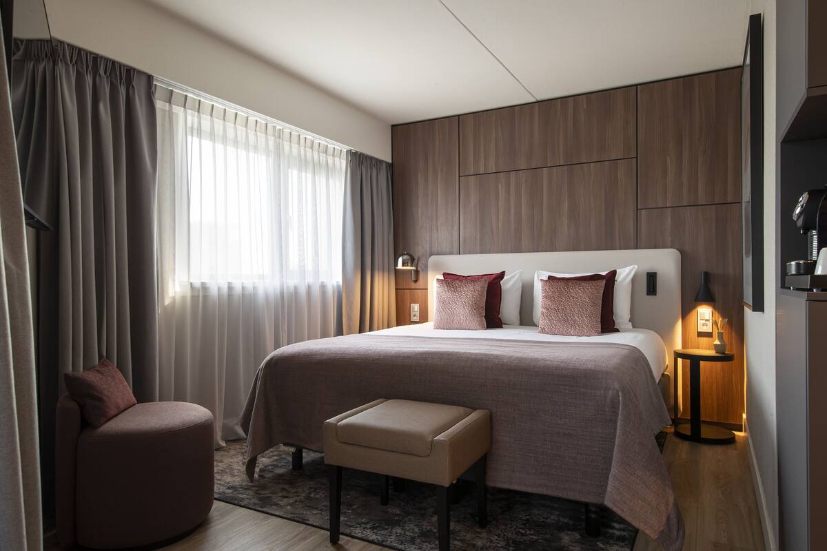 Een moderne hotelkamer met een groot bed, een nachtkastje met een lamp, een zithoek en uitzicht buiten.