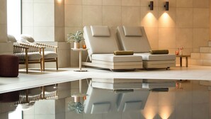 Un espace spa moderne doté d’un grand bassin aux eaux miroitantes, d’un canapé avec des coussins, d’un fauteuil en osier et d’un éclairage mural.