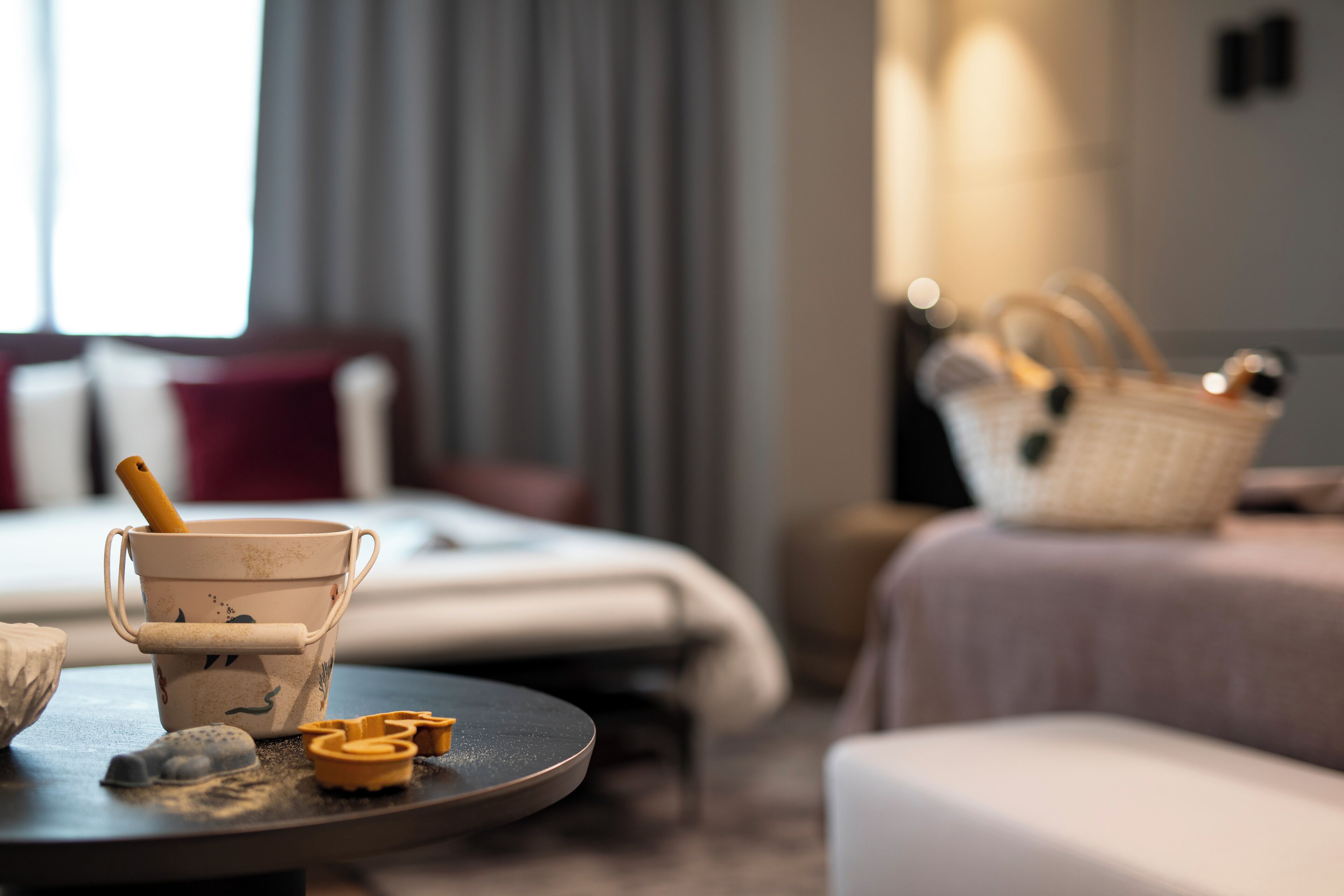 A hotel room with a bed, a table with a coffee maker, a basket, and a window with curtains.