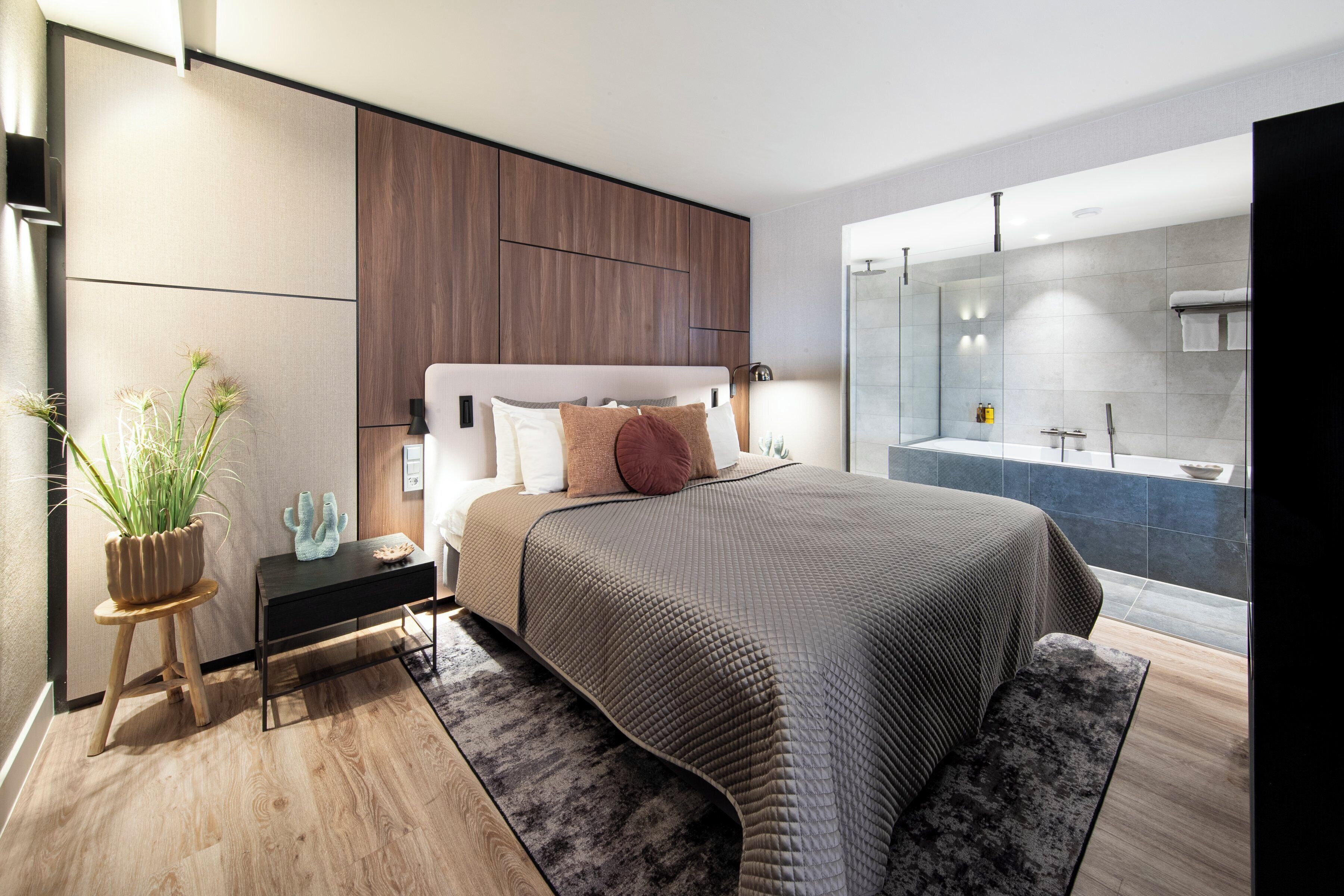 A modern bedroom with a large bed, a bedside table, a small stool with a plant, and a view of a bathroom with a bathtub and shower.