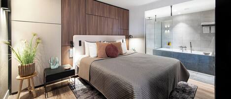 A modern bedroom with a large bed, a bedside table, a small stool with a plant, and a view of a bathroom with a bathtub and shower.