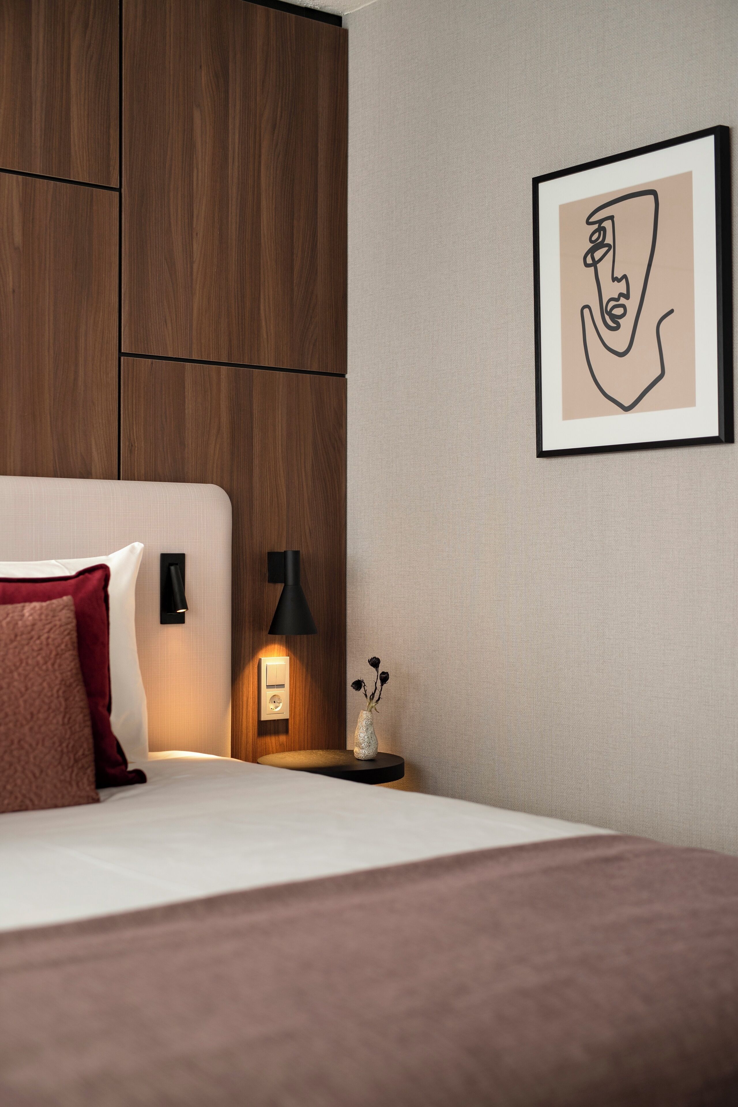 Una habitación de hotel moderna con cabecera de madera, lámpara de noche, una obra de arte enmarcada y un pequeño florero con flores sobre la mesita de noche.