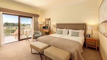 A hotel room with a large bed, a sitting area with a chair and a table, a balcony with a view, and a painting on the wall.