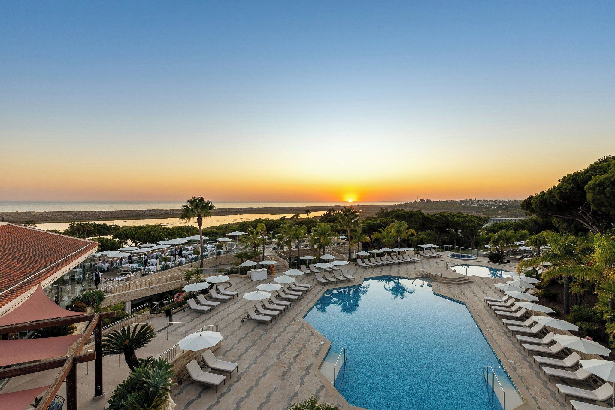 A resort with a swimming pool, sun loungers, and a view of the sunset over the sea.
