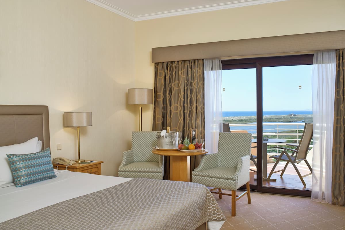 A hotel room with a bed, two chairs, a small table with a fruit bowl and a bottle, a nightstand with a phone, and a view of the ocean through a sliding door.
