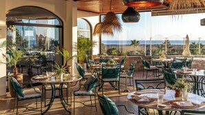 An outdoor dining area with multiple tables and chairs, surrounded by potted plants and large windows offering views of the ocean.