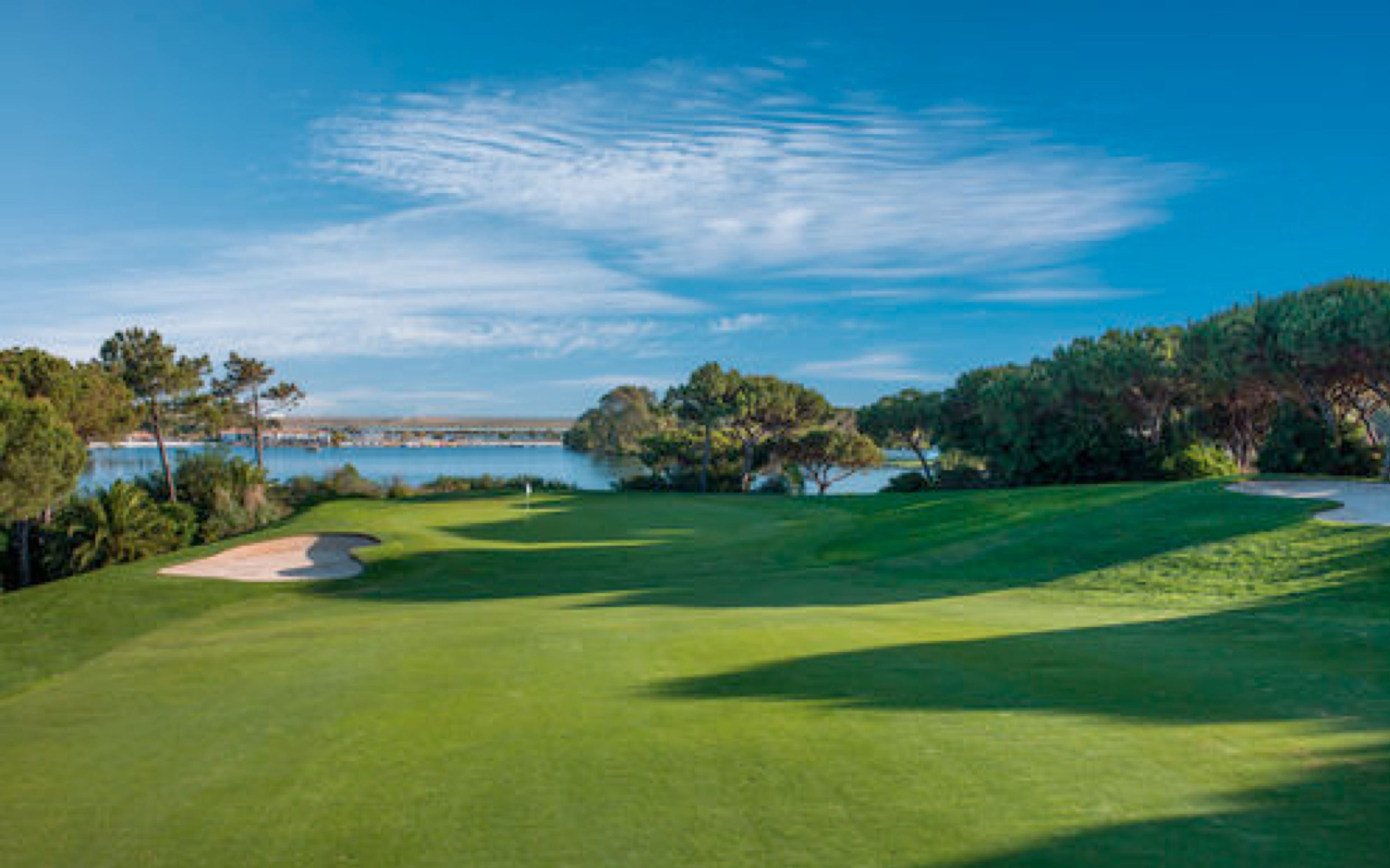 A golf course with a water body in the background, surrounded by trees and clear skies.