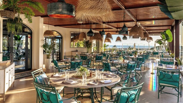 An outdoor dining area with round tables and green chairs, surrounded by potted plants and hanging thatch lamps.
