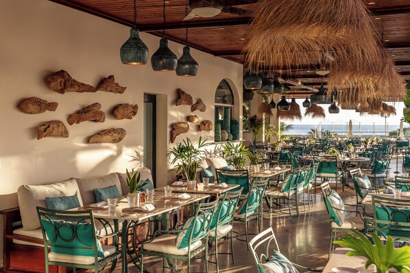 Un restaurante con vista al mar, que presenta una mezcla de decoración natural y moderna, con lámparas colgantes y esculturas de peces en la pared.
