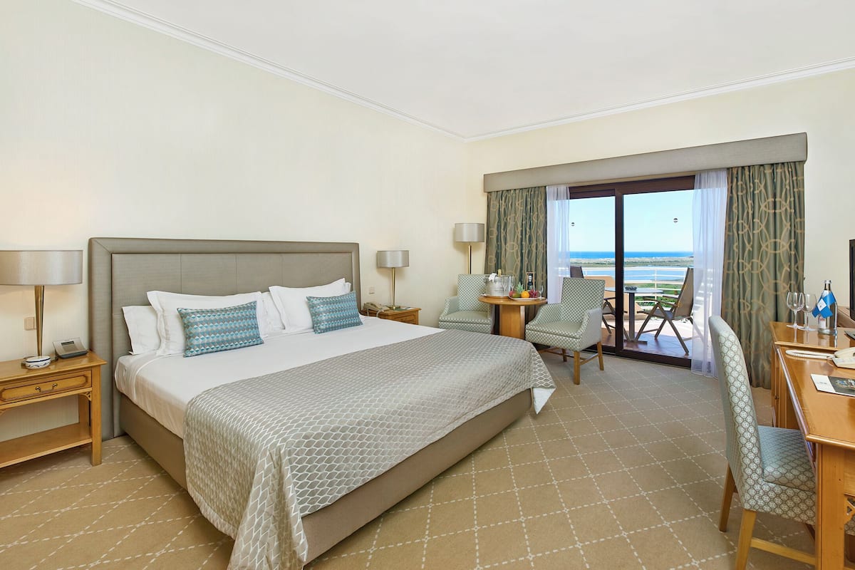 A hotel room with a large bed, a desk with a computer, a chair, a small table with a bottle and glasses, and a view of the beach through the window.