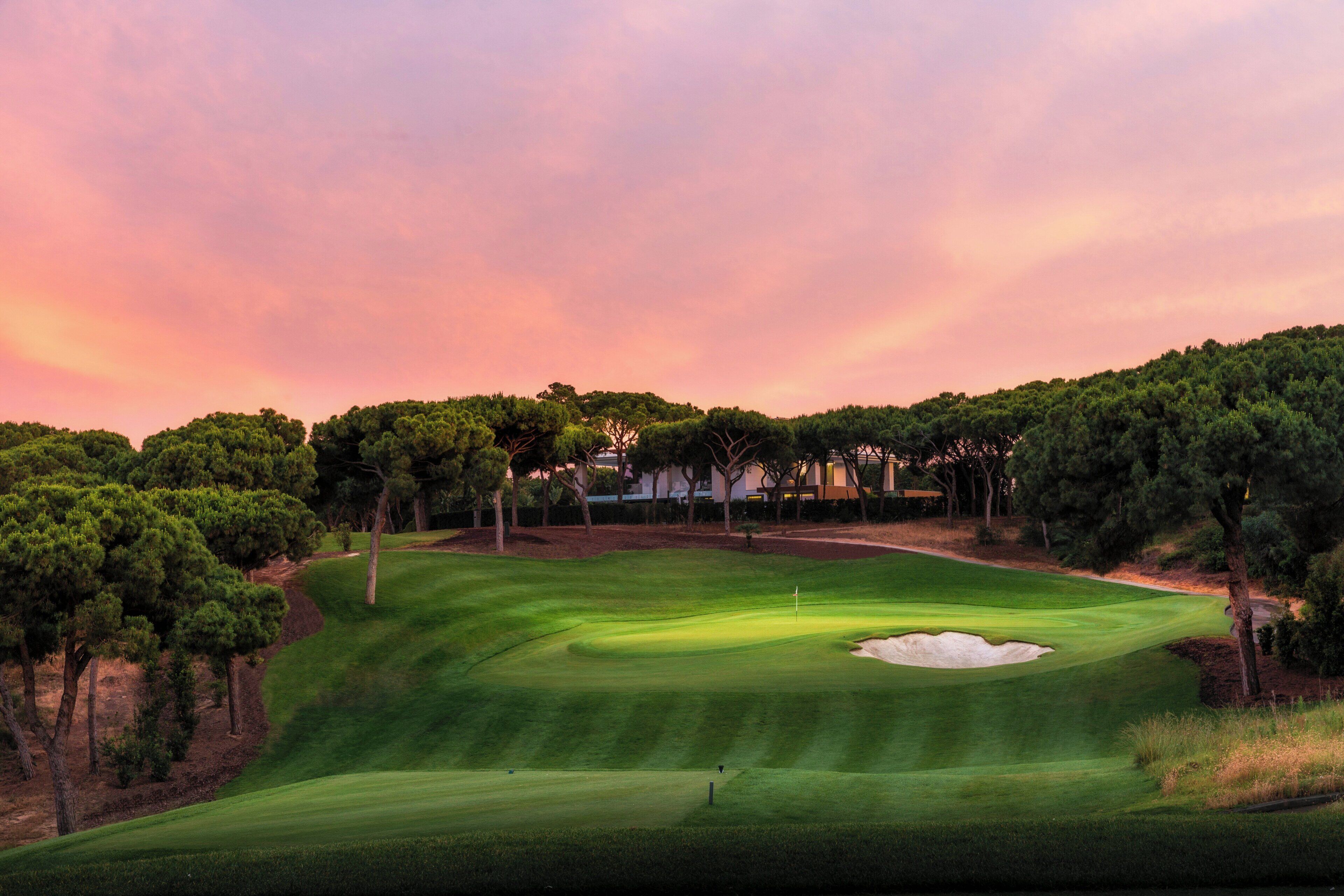 Un campo de golf con un mástil de bandera, árboles y un edificio al fondo.