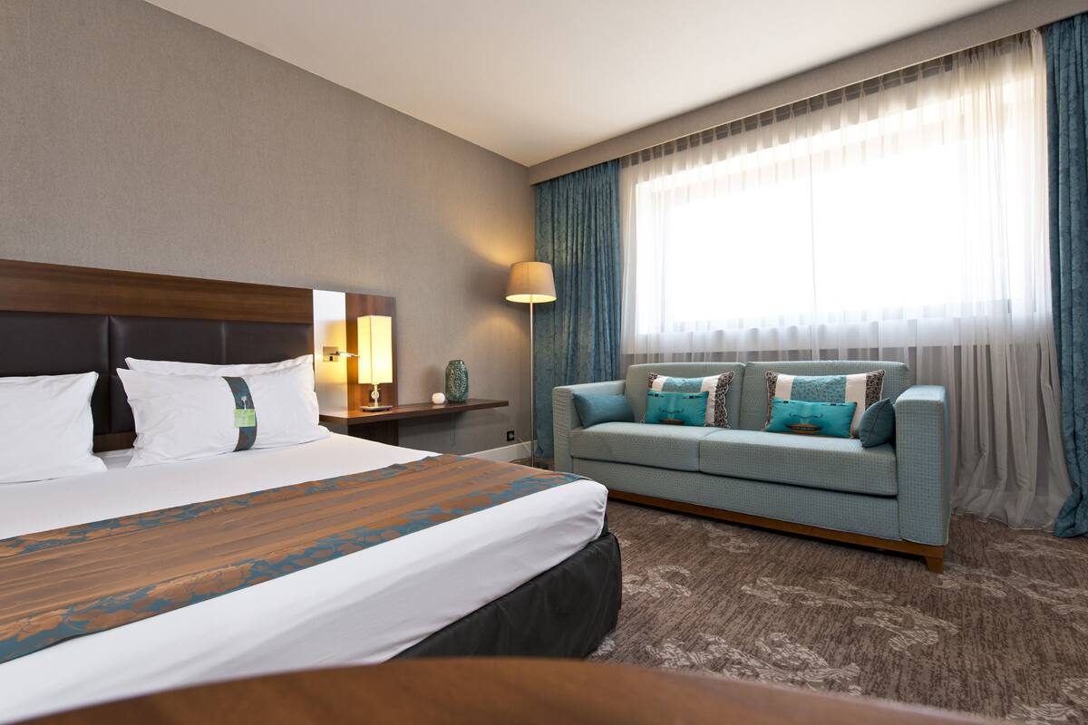 A hotel room with a large bed, a sofa, a lamp, and a window with curtains.