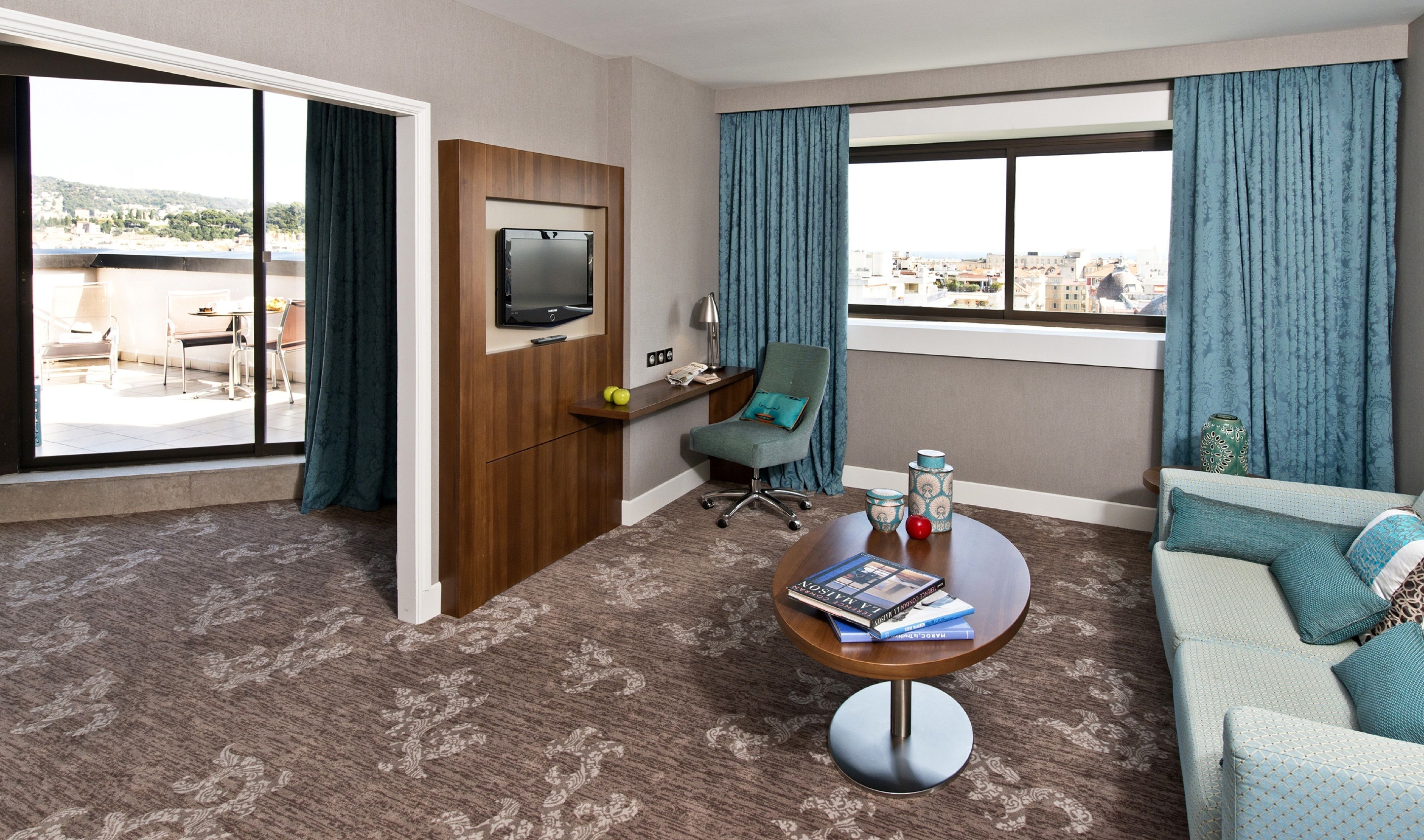 Une chambre d’hôtel dotée d’une grande fenêtre, d’un téléviseur à écran plat, d’un bureau avec une lampe, d’une chaise, d’un canapé et d’une table basse avec des livres.