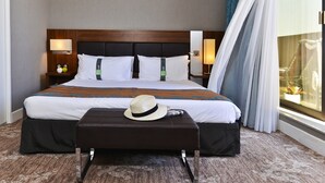 Quarto de hotel com uma cama grande, duas luminárias de cabeceira, uma mesinha de cabeceira, uma janela com cortinas e um chapéu no banquinho.