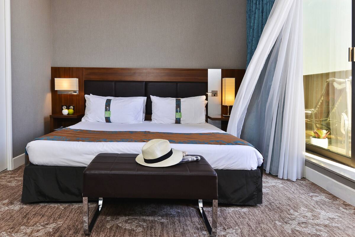 A hotel room with a large bed, two bedside lamps, a nightstand, a window with curtains, and a hat on the ottoman.