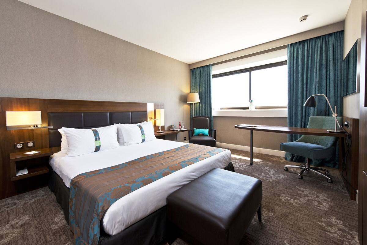 A hotel room with a large bed, a desk, a chair, a lamp, and a window with curtains.