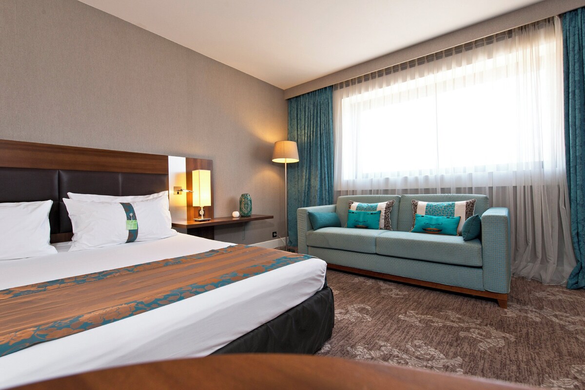 Et hotelværelse med en stor seng, en sofa, et natbord, en lampe og et vindue med gardiner.