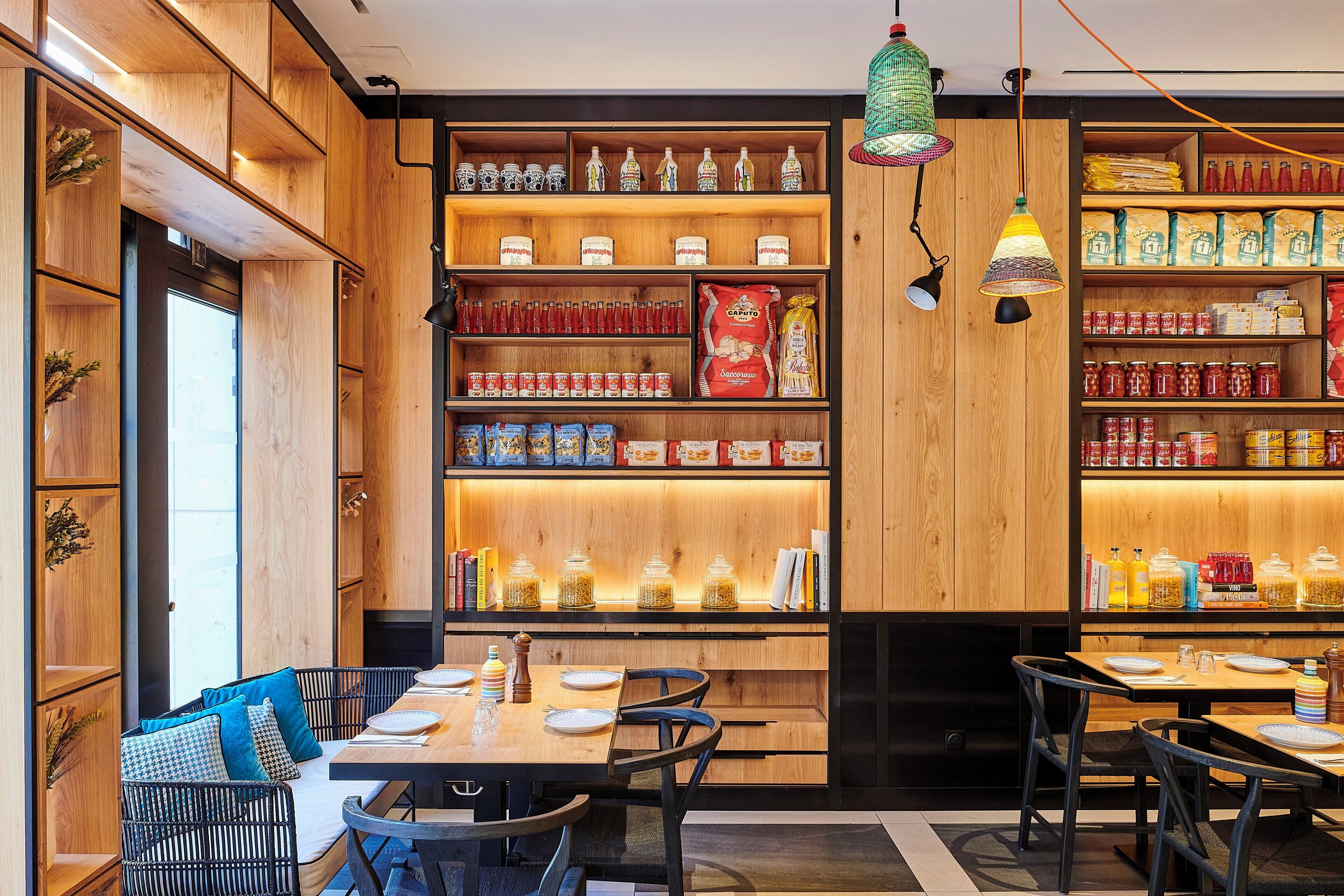 A restaurant interior with wooden shelves stocked with food items, a dining area with tables and chairs, and hanging pendant lights.