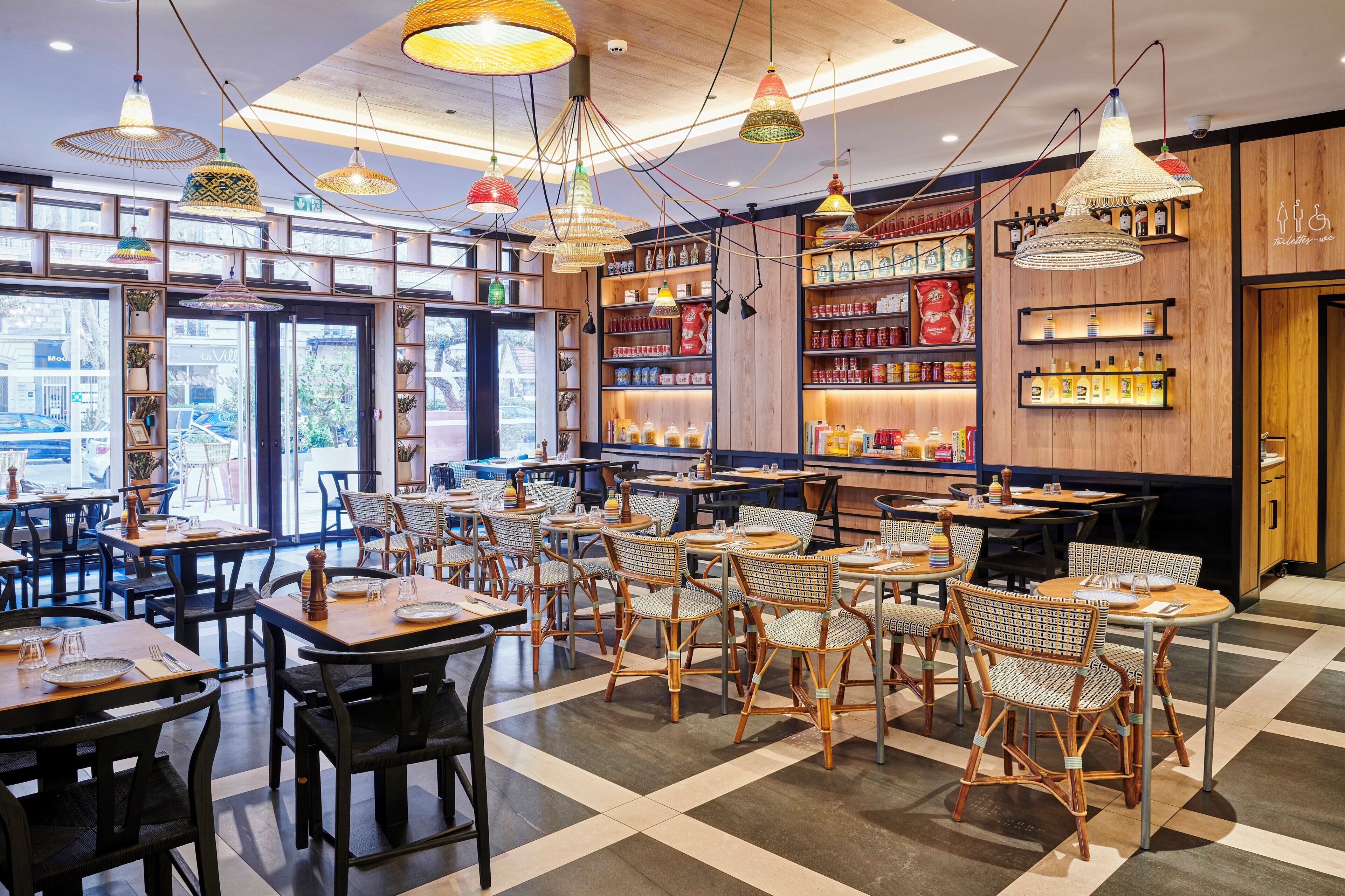 A restaurant interior with multiple dining tables and chairs, a variety of hanging pendant lights, and shelves stocked with food items.