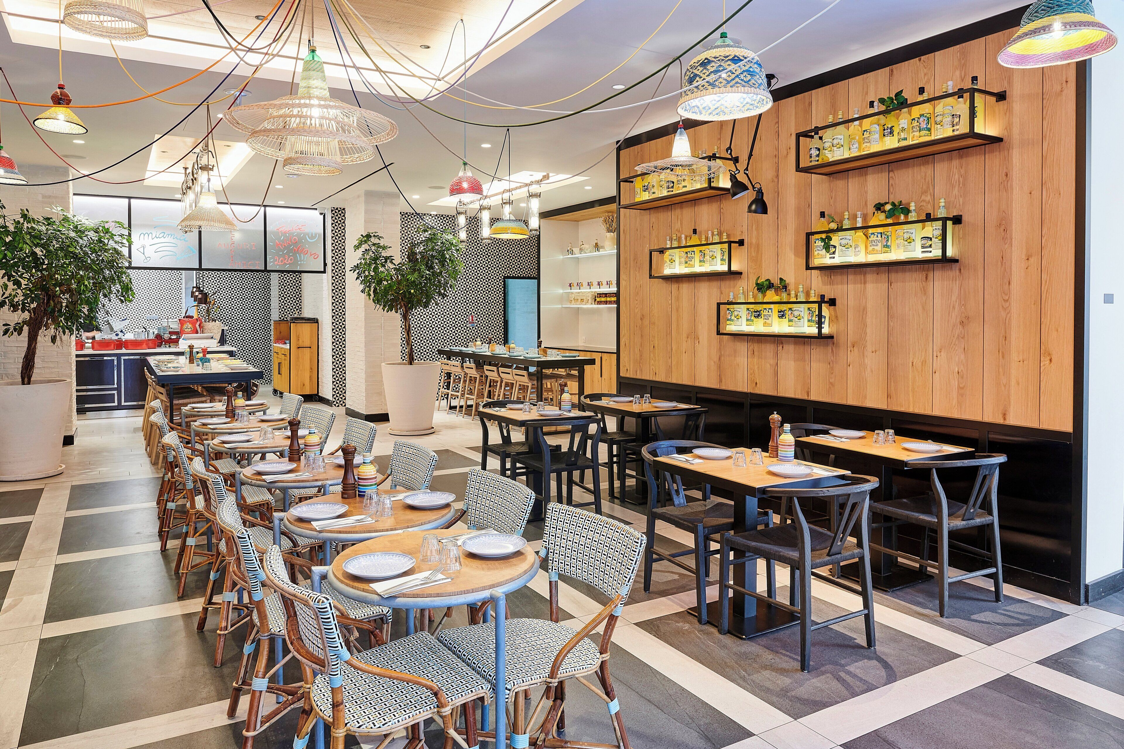 A restaurant interior with multiple dining tables and chairs, a variety of hanging lights, and shelves with bottles on the wall.